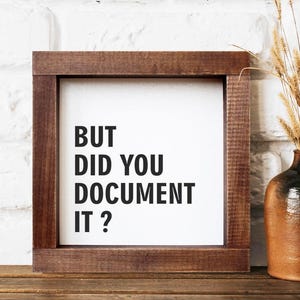Puede incluir: Un letrero enmarcado con el texto "BUT DID YOU DOCUMENT IT?" en letras negras. El marco es de color marrón oscuro. A la derecha del letrero hay un jarrón marrón y negro con espigas de trigo secas. El fondo es una pared de ladrillos blancos.