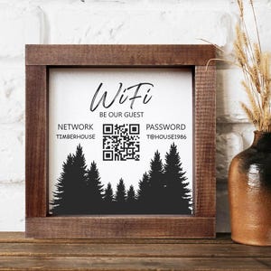 Könnte beinhalten: Ein quadratisches Holzschild mit dem Wort "WiFi" in Schreibschrift. Unter dem Wort "WiFi" steht der Satz "BE OUR GUEST". Das Schild zeigt auch den Netzwerknamen, einen QR-Code und das Passwort. Am unteren Rand befindet sich eine Silhouette von Kiefern.