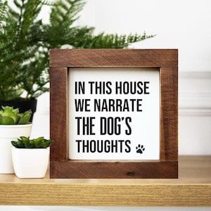 Puede incluir: Un letrero enmarcado en madera con el texto "IN THIS HOUSE WE NARRATE THE DOG'S THOUGHTS" en letras negras. Una pequeña huella de pata está en la parte inferior derecha. El letrero está sobre una estantería de madera con plantas en macetas en el fondo.