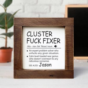 Könnte beinhalten: Ein gerahmtes Holzschild mit dem Text "CLUSTER FUCK FIXER" in schwarzer Schrift. Das Schild enthält eine Definition und den Namen "Jason" unten. Der Rahmen ist dunkelbraun.