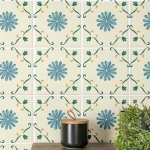 May include: A wall covered in square tiles featuring a repeating pattern of blue daisy-like flowers and green leafy vines with small orange accents on a cream background. A black cylindrical container and a green plant sit on a wooden shelf.