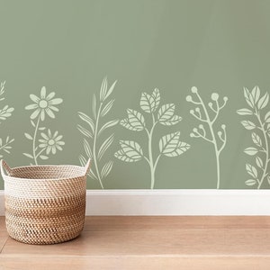 Op de afbeelding: Een geweven rieten mand staat voor een saliegroene muur met een sjabloon van witte gestileerde bloemmotieven.