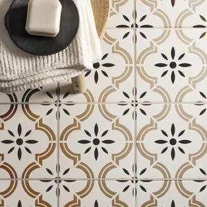 May include: Overhead shot of patterned tiles featuring a floral design in black, tan, and brown on a white backdrop. A black plate with soap and a white towel are on the left.