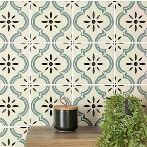 May include: Cream, teal, and black patterned ceramic tiles with a floral and geometric design. A small black jar with a wooden lid is on a wooden surface, with a potted plant in the background.