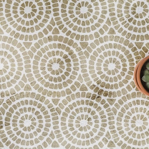 Può includere: Una superficie bianca con un motivo ripetitivo di cerchi dorati composti da piccole forme segmentate. Una parte di un vaso color terracotta contenente una pianta succulenta verde è visibile sul bordo destro dell'immagine.