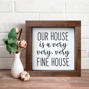 May include: A square wooden sign with the text "OUR HOUSE is a very very, very FINE HOUSE" in black script. A small white vase with greenery and cotton stems sits to the left of the sign. The sign is on a wooden surface.