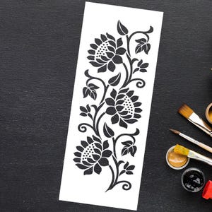 May include: A black and white floral stencil design featuring three large sunflowers with detailed petals and leaves. The stencil is on a white rectangular sheet, with paintbrushes and paint visible in the background.