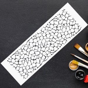 May include: A white stencil featuring a black, abstract pattern reminiscent of cracked earth or animal hide. The stencil lies on a black surface, with paintbrushes and paint pots, indicating an art or craft activity.