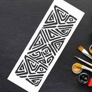 Peut inclure: Un pochoir rectangulaire blanc avec un motif géométrique noir de triangles et de lignes courbes. Le pochoir est posé sur une surface noire, avec des pinceaux et des pots de peinture, suggérant un projet artistique.