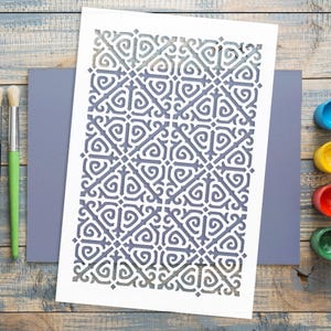 May include: A white stencil featuring a repeating geometric design, placed on a grey surface. A green-handled paintbrush and small pots of blue, yellow, red, and green paint are on a wooden surface.