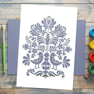 Può includere: Una carta bianca con un disegno floreale e di uccelli grigio, posta su una busta grigia. Il disegno presenta un vaso con due uccelli e un motivo floreale. Pennelli e barattoli di vernice sono visibili su una superficie di legno.