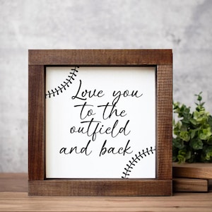 Puede incluir: Un letrero cuadrado de madera con un marco marrón oscuro. El letrero tiene un fondo blanco con el texto "Love you to the outfield and back" en escritura negra. Los diseños de costura de béisbol están en las esquinas.