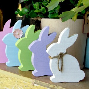 May include: Five wooden bunny figurines in a row, painted in pastel colors: pink, blue, green, purple, and white. The bunnies are arranged from left to right in order of color.
