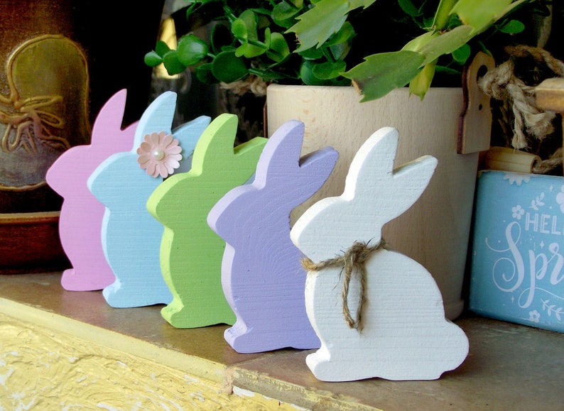 May include: Five wooden bunny figurines in a row, painted in pastel colors: pink, blue, green, purple, and white. The bunnies are arranged from left to right in order of color.
