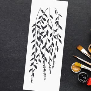 May include: A black and white stencil of a weeping willow branch design. The stencil is on a white rectangular surface, with paintbrushes and paint pots in the background. The design features leaves and hanging branches.