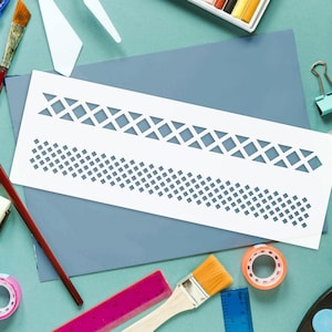 Pode incluir: Um estêncil branco com padrões geométricos, incluindo losangos e quadrados, está sobre papel azul. Materiais de arte como pincéis, fita adesiva, réguas e pastéis circundam o estêncil, criando uma cena de espaço de trabalho criativo.