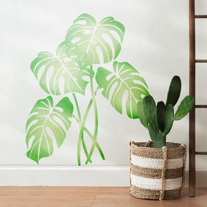 Op de afbeelding: Een aquarel van een tropische plant met grote groene bladeren. De plant is geschilderd op een witte muur en is omgeven door een witte rand. Een kleine cactusplant staat in een geweven mand in de rechterbenedenhoek van de afbeelding.