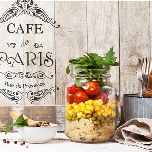 May include: A mason jar filled with a layered salad with quinoa, peas, corn, tomatoes, and fresh herbs. The jar is on a white wooden surface with a sign that reads "Cafe de Paris Rue de Provence".