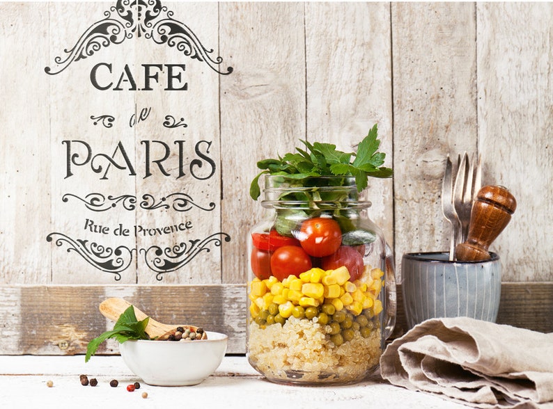 May include: A mason jar filled with a layered salad with quinoa, peas, corn, tomatoes, and fresh herbs. The jar is on a white wooden surface with a sign that reads "Cafe de Paris Rue de Provence".