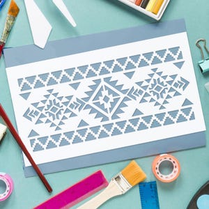May include: A white stencil with a geometric pattern, featuring triangles and diamond shapes, is placed on a blue surface. The stencil is surrounded by art supplies, including paintbrushes, tape, and rulers, suggesting a craft or DIY project.