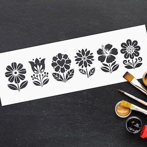 Op de afbeelding: Een witte papieren strook met zes zwarte bloempatronen. De ontwerpen omvatten verschillende bloemsoorten, zoals madeliefjes, tulpen en hibiscus. Penselen en verfpotten met gele en zwarte verf liggen op een zwart oppervlak.