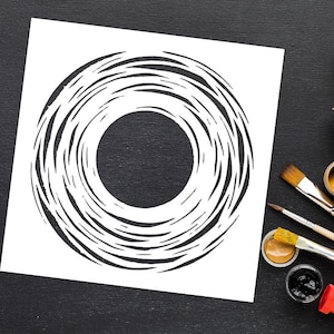 May include: A white stencil with a circular design, a central hole, and swirling lines. The stencil is on a black surface. Paintbrushes and paint pots are in the corner, indicating an art or craft project.