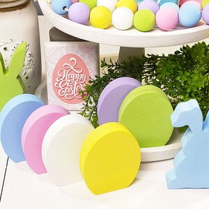 May include: A set of six pastel-colored wooden eggs and two wooden bunny figurines. The eggs are arranged in a row, with the bunny figurines on either end. The eggs are painted in shades of blue, pink, white, and yellow. The bunnies are painted in shades of green and blue. The eggs and bunnies are all decorated with a simple, rustic style.