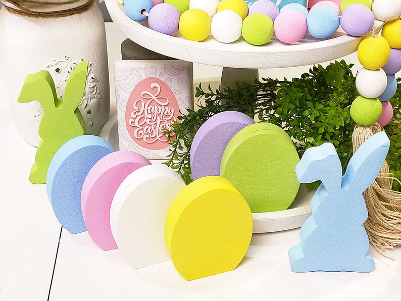 May include: A set of six pastel-colored wooden eggs and two wooden bunny figurines. The eggs are arranged in a row, with the bunny figurines on either end. The eggs are painted in shades of blue, pink, white, and yellow. The bunnies are painted in shades of green and blue. The eggs and bunnies are all decorated with a simple, rustic style.