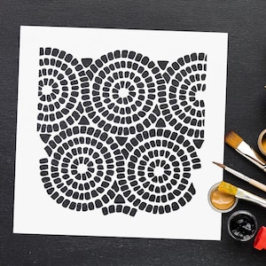 May include: A black and white stencil design featuring a repeating pattern of concentric circles. The stencil is on a white sheet of paper, with paintbrushes and paint pots in the background, suggesting an art or craft project.