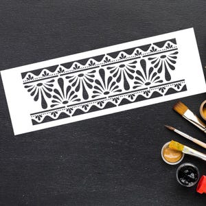 May include: A white stencil with a repeating black floral design, featuring stylized flower-like shapes and dotted lines. The stencil is on a black surface, with paintbrushes and paint pots in the corner, suggesting its use in art or craft projects.