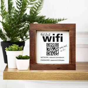 Könnte beinhalten: Ein Holzschild mit den Worten "But first WIFI" in Schwarz, einem QR-Code sowie Netzwerk- und Passwortdetails. Das Schild steht auf einem Holzregal mit Topfpflanzen.