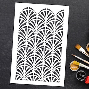 May include: A white stencil featuring a repeating Art Deco fan pattern in black. The stencil is on a white surface, with paintbrushes and paint pots visible on a black surface in the background. This stencil is ideal for DIY home decor projects.