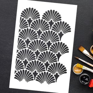 May include: A black and white stencil featuring a repeating fan-like pattern. The design is on a white background, with paintbrushes and paint pots visible on a black surface, suggesting its use in art or craft projects.