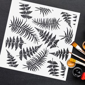 May include: A white stencil sheet with a repeating pattern of fern leaves. The stencil is on a black surface with paint brushes and paint jars.