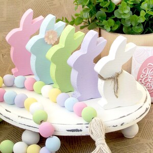 May include: A white tiered tray with pastel colored wooden bunny figurines and a pastel colored beaded garland. The tray has a distressed white finish and a sign that says "Happy Easter".