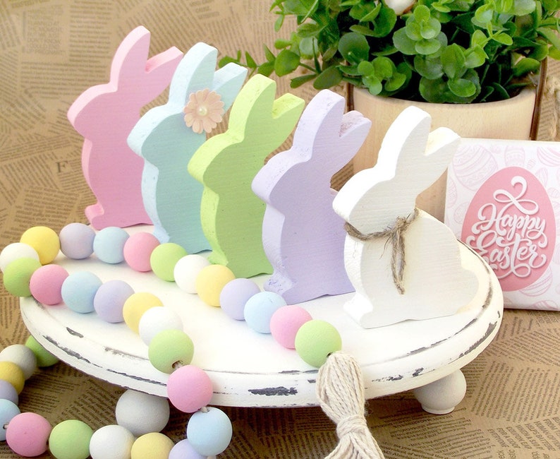 May include: A white tiered tray with pastel colored wooden bunny figurines and a pastel colored beaded garland. The tray has a distressed white finish and a sign that says "Happy Easter".