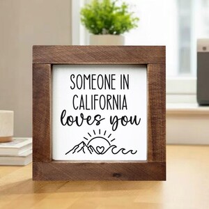 以下が含まれることがあります： 「SOMEONE IN CALIFORNIA loves you」という黒い文字が書かれた正方形の木製サイン。山、太陽、ハート、波のデザインが特徴です。フレームは濃い茶色の木材で作られています。