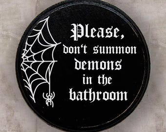 May include: Black round wooden sign with white text that reads "Please, don't summon demons in the bathroom". The sign has a white spider web and spider graphic.