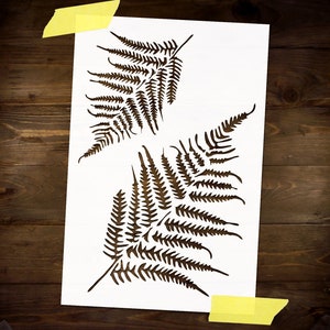 May include: A white stencil with two fern leaf designs cut out. The stencil is on a brown wooden surface.