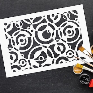 May include: A white stencil with a black circular design of varying sizes and concentric circles. The stencil is on a black surface, with paintbrushes and paint pots nearby, suggesting an art or craft project.
