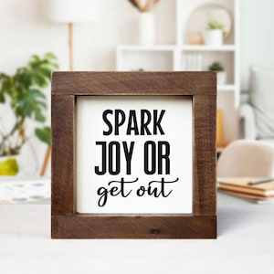以下が含まれることがあります： 黒い文字で「SPARK JOY OR get out」と書かれた正方形の木製サイン。サインは白い背景と濃い茶色の木製フレームです。サインは白い表面に置かれています。