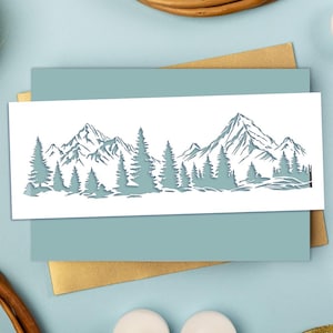 May include: A white rectangular stencil featuring a detailed laser-cut landscape design. The artwork displays a panoramic mountain range with jagged peaks in the background and a dense forest of evergreen pine trees in the foreground.
