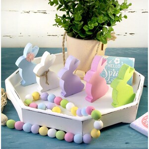May include: A white tray with a pastel colored beaded garland and five wooden bunny figurines in shades of blue, white, purple, pink, and green. The bunnies have a flower detail on their heads. The tray has a sign that says "Hello Spring" on it.