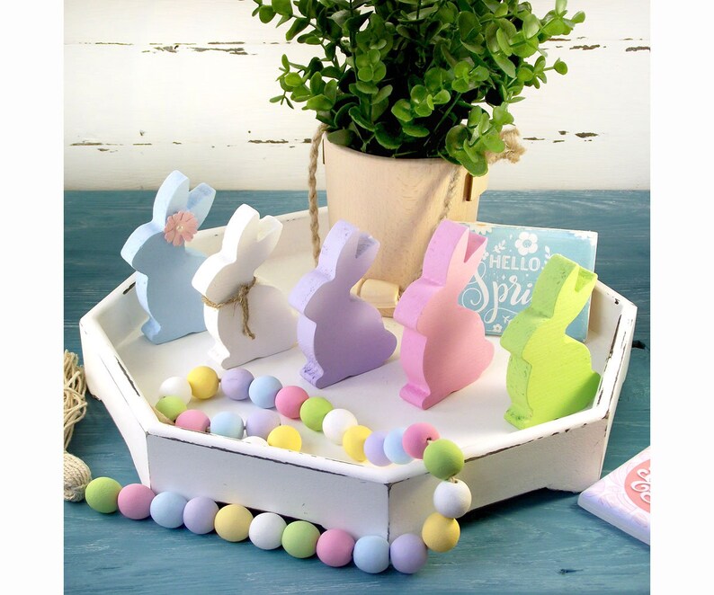 May include: A white tray with a pastel colored beaded garland and five wooden bunny figurines in shades of blue, white, purple, pink, and green. The bunnies have a flower detail on their heads. The tray has a sign that says "Hello Spring" on it.