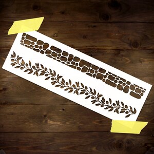 May include: White stencil with two designs. The top design is a row of stones. The bottom design is a row of olive branches with leaves and berries.