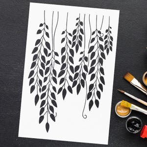 May include: Black and white botanical art print featuring stylized hanging leaves. The artwork is on a white background, with paintbrushes and paint pots visible on a black surface. The leaves are detailed with a minimalist design.