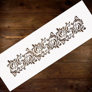 May include: White stencil with a decorative floral design. The design is a repeating pattern of leaves and swirls. The stencil is on a brown wooden surface.