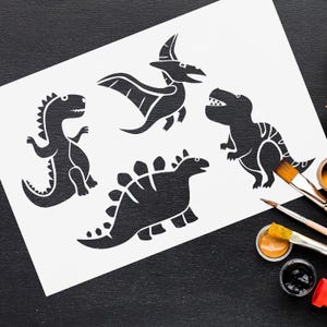 May include: A white sheet of paper with four black dinosaur illustrations. The dinosaurs are in various poses, with one having wings. Paintbrushes and paint pots with brown and black paint are on a black surface.
