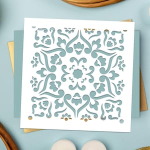 May include: A white square stencil featuring a symmetrical, ornate design in a light teal hue. The design incorporates floral and scroll-like motifs. The stencil rests on a light teal backdrop with gold and white accents, indicating a craft or DIY project.
