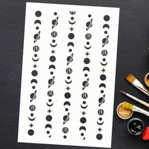 May include: A white stencil sheet with repeating patterns of celestial bodies. The design includes planets with rings, crescent moons, stars, and circles. The sheet is on a black surface with paintbrushes and paint pots.